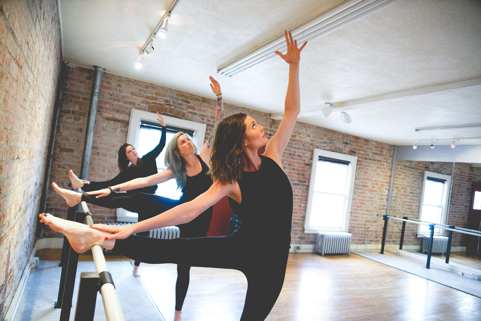 Three women doing a barre workout with arms stretched upward.