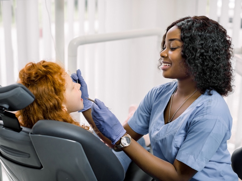 Side view of curly red haired girl lying on dentist chair with open mouth while female African doctor in blue uniform makes dental checkup with tools. Concept of teeth treatment and care.