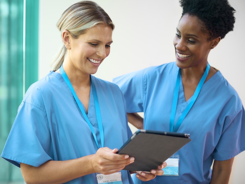 Two Mature Female Dentists Wearing Scrubs With Digital Tablet Discussing Dental Patient Scheduling